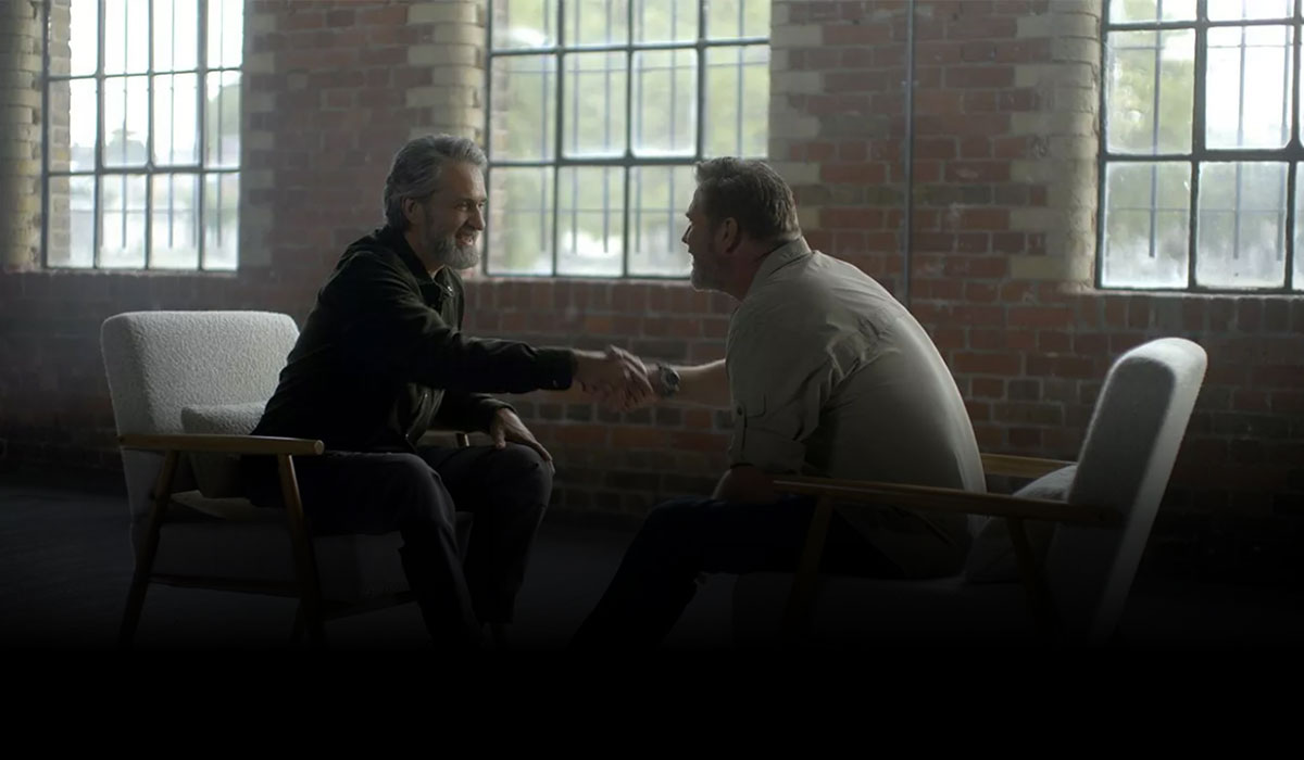 A photograph depicts two men engaged in a handshake, sitting across from each other in a room that is filled with natural light from large windows. The man on the left, who is identified as Lawrence Gunther, has a full head of graying hair, a neatly groomed beard, and is smiling warmly while extending his hand. He is dressed in a casual green jacket over a dark shirt. The room has an industrial feel, with a high ceiling, exposed brick walls, and a clear view of the sky through the windows. The man on the right, Brent Stirton, is sitting with his back slightly turned towards the camera, which obscures a full view of his features. He is wearing a light-colored, long-sleeve shirt and seems to be reciprocating the handshake. The atmosphere is friendly and open. There are two chairs in the scene: the one on the left is a mid-century modern style with a white cushion, while the one on the right has a minimalistic design, blending with the light and airy ambiance of the setting.