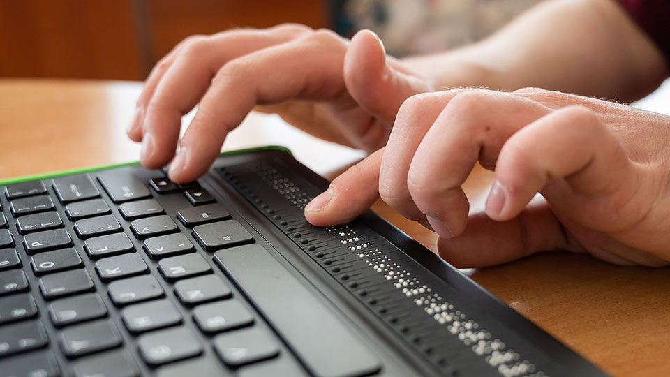 This image shows a close-up of a person's hands using a computer keyboard and a refreshable braille display. The braille display is positioned below the keyboard and features a row of small, raised dots that can be felt with the fingers to read braille text. The fingers of the person are actively engaged with both the keyboard and the braille display, indicating they are typing and reading simultaneously. The braille display consists of tactile bumps that change dynamically to represent different characters in braille. This setup is commonly used by visually impaired individuals to interact with digital devices.